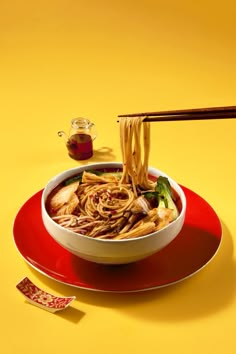 a bowl of noodles and chopsticks on a red plate with a yellow background