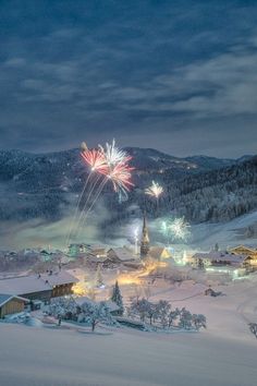 fireworks are lit up in the sky above a snow covered village and mountains at night