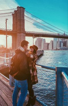 a man and woman standing next to each other on a pier near the water with a bridge in the background