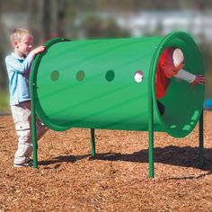 a little boy playing in a green play structure on the ground with another child standing next to it