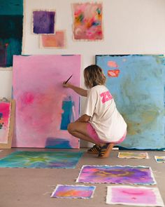 a woman kneeling down in front of paintings on the floor and holding a paintbrush