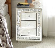 a mirrored night stand with three drawers and two mirrors on the top, in front of a white bed