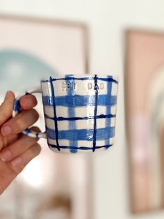 a hand holding a blue and white striped coffee cup in front of a wall with pictures on it