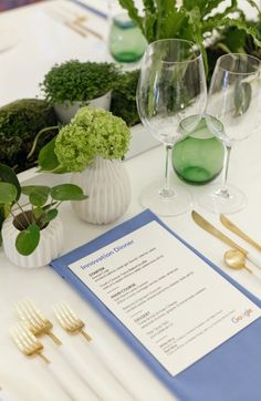 the table is set with glasses, silverware and green plants in white vases