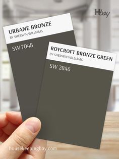 The image compares two Sherwin Williams paint swatches. The left swatch is labeled "Urbane Bronze" with the code SW 7048, a dark, rich brown with gray undertones. The right swatch is "Roycroft Bronze Green" with the code SW 2846, a deep, muted green with bronze undertones. Both swatches are held by a hand, with a blurred indoor background in neutral tones. These colors are bold and versatile, suitable for accent walls or exterior use. Urbane Bronze Sherwin Williams Kitchen Cabinets, Roycroft Bronze Green Sherwin Williams Exterior, Sherwin Williams Deep Green, Sw Urbane Bronze Accent Walls, Urban Bronze Exterior, Roycroft Bronze Green Sherwin Williams, Roycroft Bronze Green, Trim Color Ideas, Sw Urbane Bronze