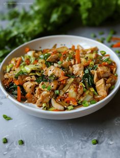 a white bowl filled with chicken and vegetables on top of a gray table next to lettuce