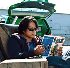 a woman reading a book while sitting in a chair next to a green pickup truck