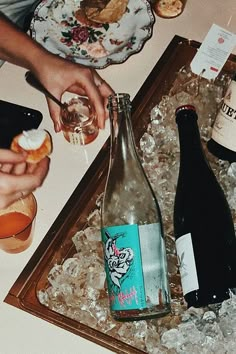 two people holding wine glasses near bottles and ice on a table with other food items