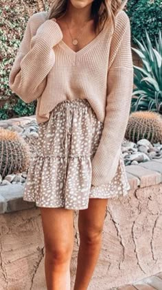 a woman wearing a tan sweater and polka dot skirt, standing in front of a cactus