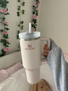 a person holding a coffee cup with a teddy bear on the bed in front of it