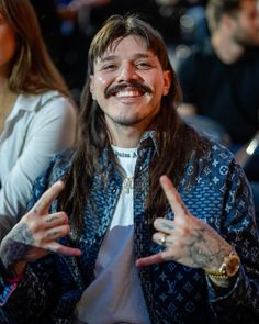 a man with long hair and tattoos sitting in front of a crowd giving the peace sign