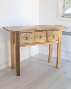 a wooden table with three drawers on it in a white room next to a window