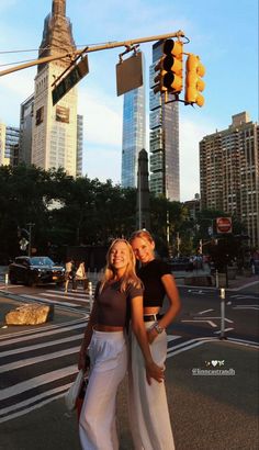 two women are standing in the middle of an intersection with traffic lights and skyscrapers behind them