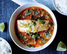 a bowl filled with fish and vegetables next to rice on a blue table cloth, surrounded by lime wedges