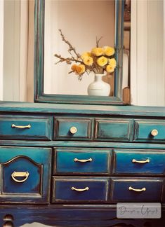 a blue dresser with yellow flowers in a vase on top and a mirror above it