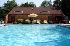 an empty swimming pool in front of a brown building with umbrellas and chairs around it