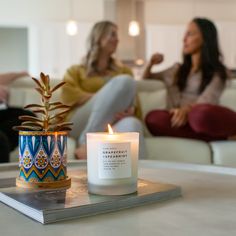 two women sitting on a couch in the background, one is holding a candle and the other has a potted plant