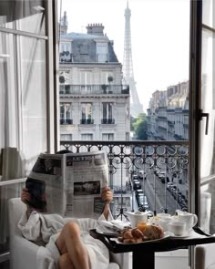 a woman sitting in a chair reading a newspaper while holding a cup of coffee and looking at the eiffel tower