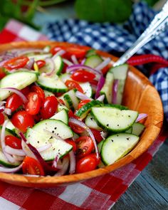 a wooden bowl filled with cucumbers, tomatoes and onions on top of a checkered table cloth