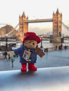 a teddy bear with a red hat and blue coat is standing in front of the london bridge