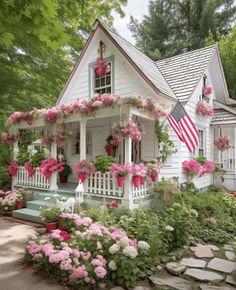 a white house with pink flowers and an american flag hanging from the front porch, surrounded by greenery