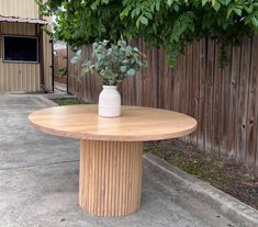 a wooden table with a white vase on top and green plants in the middle, outside