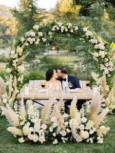 a bride and groom sitting at a table in front of an arch with white flowers