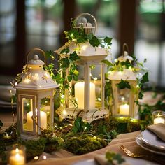 three white lanterns with lit candles are sitting on a table surrounded by greenery and place settings