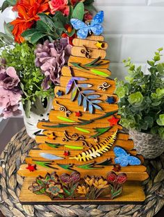 a wooden christmas tree with colorful flowers and butterflies on it's top, next to a potted plant