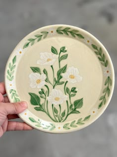 a hand holding a white and green plate with flowers painted on the side, in front of a gray background