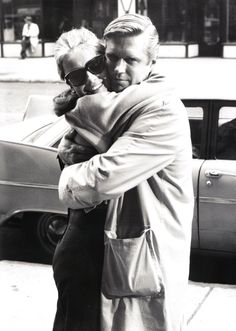 an old man and woman hugging on the street in front of a car, black and white photograph