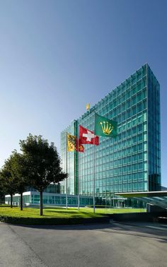 a large glass building with flags flying in the wind next to a tree and grass area