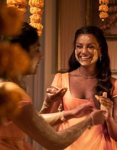 a woman in an orange dress smiles as another woman holds something up to her face