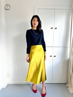 a woman standing in front of a white wall wearing a black sweater and yellow skirt