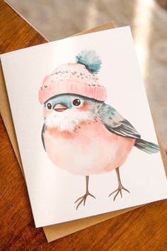 a card with a watercolor painting of a bird wearing a pink knitted hat