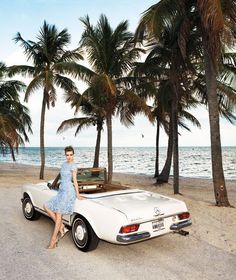 a woman sitting on the back of a white car near some palm trees and water