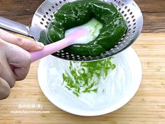 a person using a strainer to scoop green vegetables into a white bowl on top of a wooden table