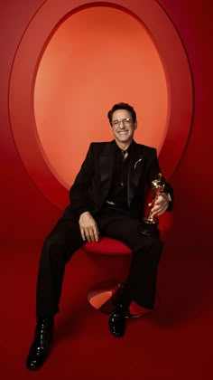 a man in a black suit holding an award sitting on a red chair with a circular background