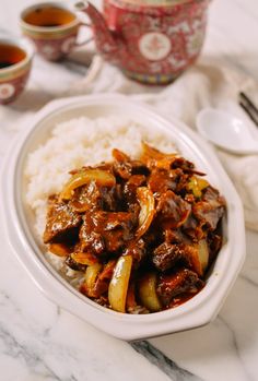 a white bowl filled with meat and vegetables on top of rice next to two cups of tea