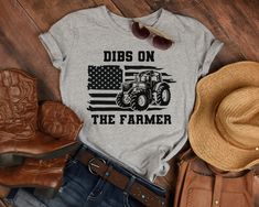 "This shirt is the perfect addition to your wardrobe for the farm-loving, country-chic enthusiast. This comfortable and stylish t-shirt features a bold and eye-catching graphic of an American flag and a tractor, surrounded by text that reads \"Dibs on the Farmer\". This t-shirt is ideal for all-day wear, whether you're out on the farm or running errands in town.  The classic fit and crew neck make it easy to wear, while the bold graphic adds a touch of personality and flair. Whether you're a far Farm Wife Shirt, Farm Wife, Farmer Shirt, The Farmer, A Farmer, On The Farm, Christmas 2024, Country Chic, The Farm