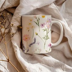 a coffee cup with flowers painted on it sitting on a white sheet next to dried plants