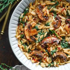 pasta with mushrooms and spinach in a white bowl on a table next to gold utensils