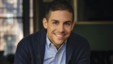 a smiling man wearing a blue sweater and cardigan sitting in front of a window