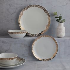 three white and gold dinner plates on a marble countertop next to a vase with flowers