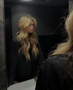 a woman standing in front of a mirror looking at her long blonde hair and wearing a black top