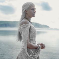 a woman with long white hair is standing by the water and looking off into the distance