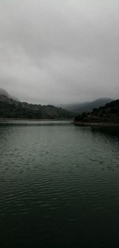 a body of water surrounded by hills and clouds in the distance on a cloudy day