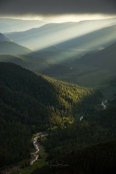 the sun shines through the clouds in the mountains above a valley with a river running through it