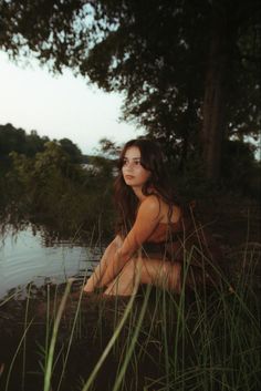 a woman is sitting in the water by some tall grass and looking off into the distance