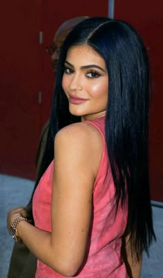 a woman with long black hair wearing a pink top and bracelets standing in front of a red wall